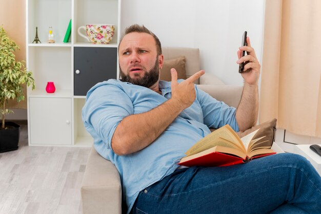 Clueless adult slavic man sits on armchair holding book on legs pointing at phone and looking at side inside the living room