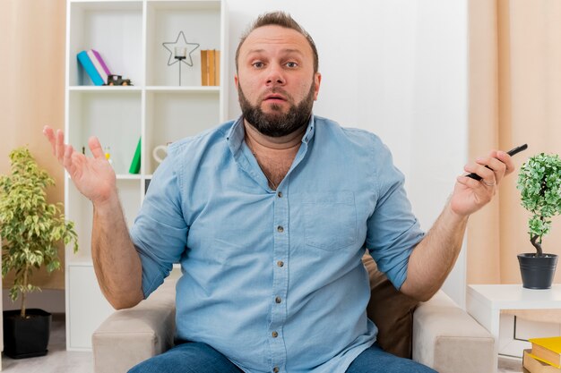 Clueless adult slavic man sits on armchair holding tv remote inside the living room