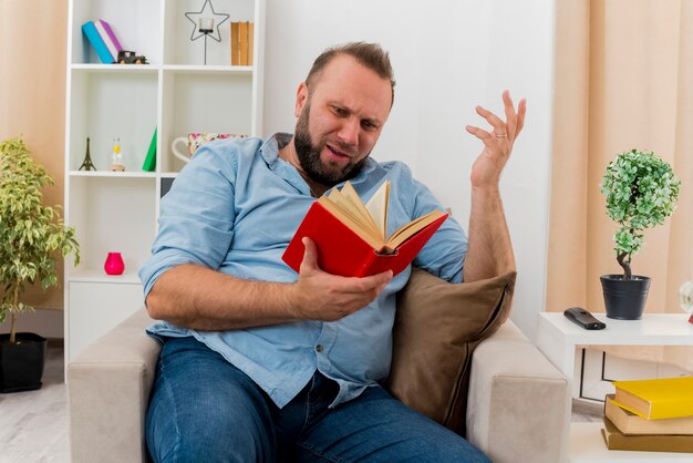 Clueless adult slavic man sits on armchair raising hand holding and looking at book inside the living room