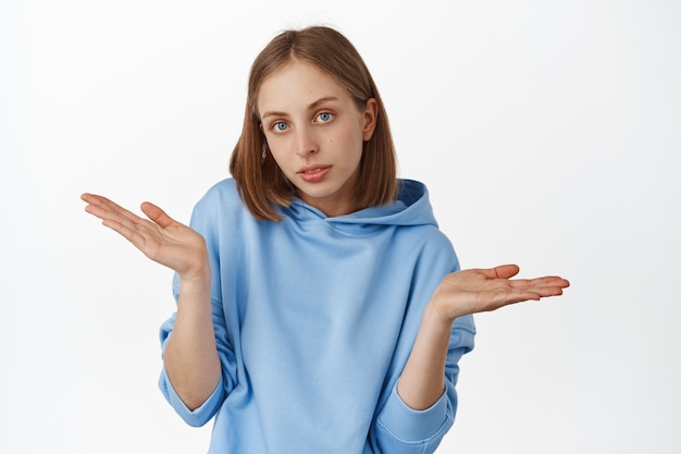 Free photo clueless blond caucasian woman shrugging shoulders, spread empty hands and look unaware, indecisive what to do, standing over white wall.