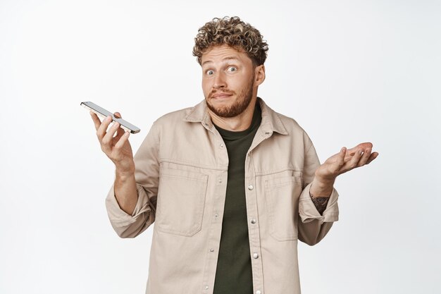 Clueless blond guy shrugging shoulders holding smartphone and looking unaware dont know anything have nothing to say standing over white background