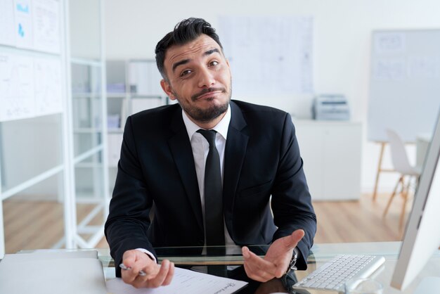 Clueless Caucasian businessman sitting at desk in office and looking at camera