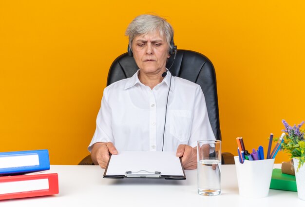 Clueless caucasian female call center operator on headphones sitting at desk with office tools holding and looking at clipboard