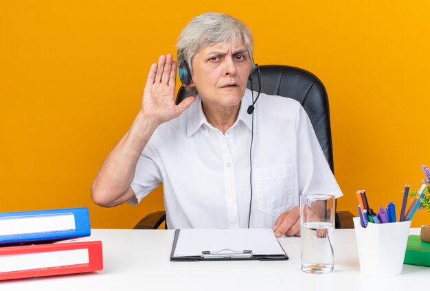 Clueless caucasian female call center operator on headphones sitting at desk with office tools keeping hand close to her ear trying to hear isolated on orange wall