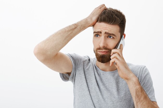 Clueless concerned young man with beard and moustache feeling uneasy and uncertain making decision talking on smartphone scratching head and frowning with sad expression, frustrated over white wall