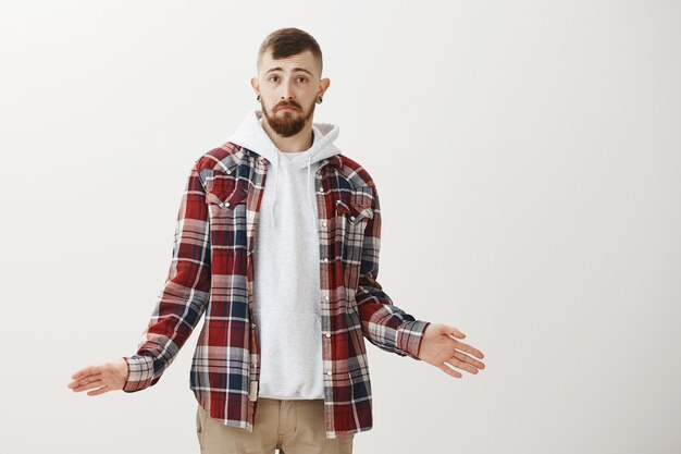 Clueless and confused hipster guy shrugging and spread hands sideways unaware