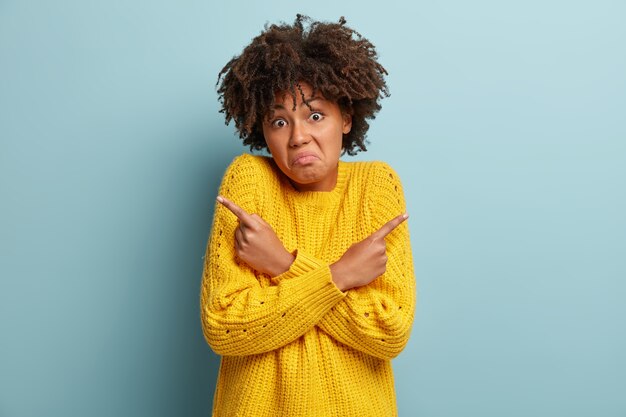 Clueless confused woman points at different corners, feels doubtful and unaware, makes decision, wears knitted yellow jumper, frowns face, stands over blue wall. People and perception concept