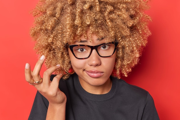 Clueless curly haired young woman shrugs shoulders with bewilderment has hesitant face expression cannot make decision wears transparent glasses and black t shirt isolated over vivid red background