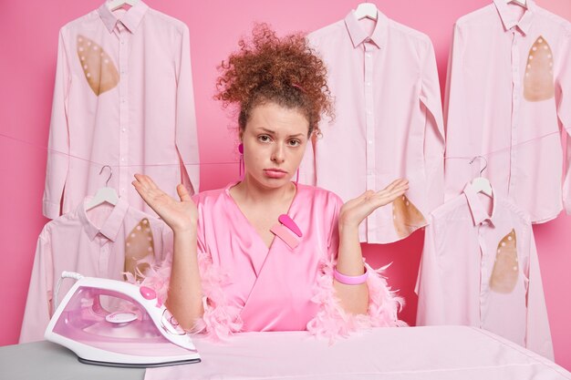 Clueless curly woman spreads palms feels hesitant does housework wears dressing gown irons folded clothes isolated over pink wall doesnt know what to do. Doubtful housewife near ironing board