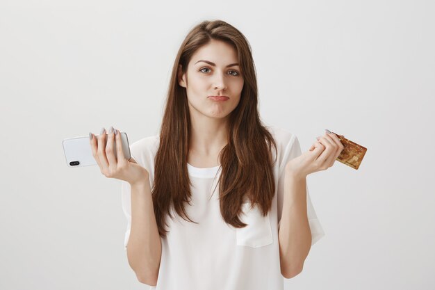 Clueless cute girl shrugging with credit card and mobile phone