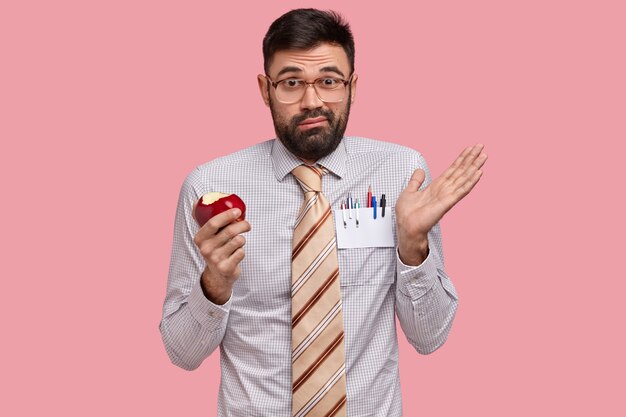 Clueless discontent bearded man spreads hand, holds red apple, wears optical glasses and formal clothes, feels doubt