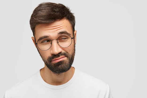 Foto gratuita un uomo dubbioso senza tracce con una folta barba scura, guarda con esitazione a parte, alza le sopracciglia perplesso