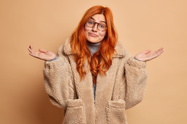 Clueless doubtful redhead young Caucasian woman spreads palms and faces difficult choice wears brown fur coat gestures indecisive.