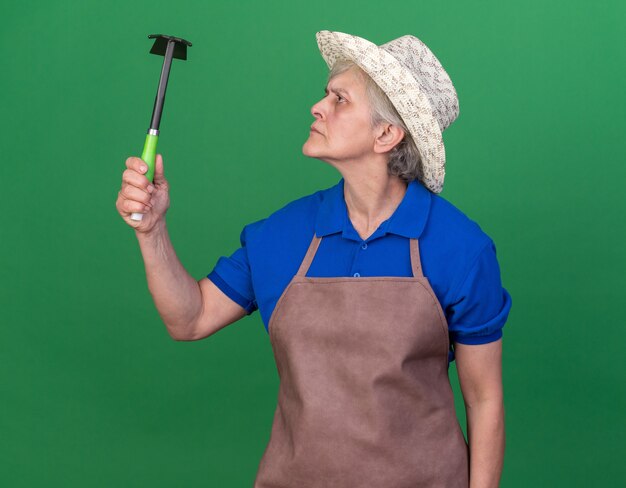 Clueless elderly female gardener wearing gardening hat holding and looking at hoe rake