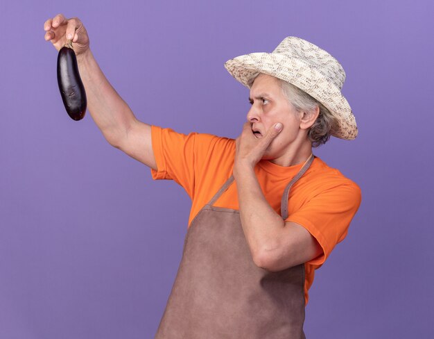 Бестолковая пожилая женщина-садовник в садовой шляпе кладет руку на подбородок и смотрит на баклажаны на фиолетовом