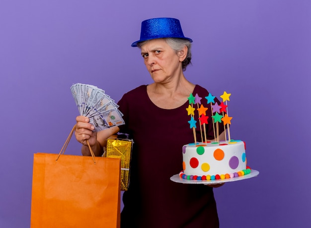パーティーハットを身に着けている無知な年配の女性は、コピースペースで紫色の壁に分離されたお金のギフトボックス紙の買い物袋とバースデーケーキを保持します