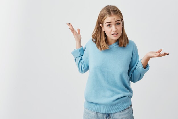 Clueless girl dressed in sweater and jeans shrugging shoulders, staring  with confused look after she did something wrong, not feeling sorry or guilty, not understanding what is wrong.