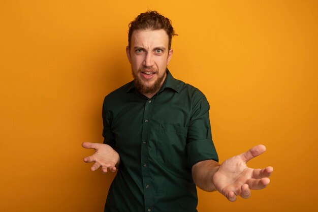 Clueless handsome blonde man holds hands open looking at camera on orange