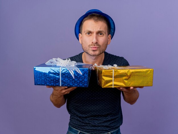 Clueless handsome caucasian man wearing blue hat holds gift boxes isolated on purple background with copy space