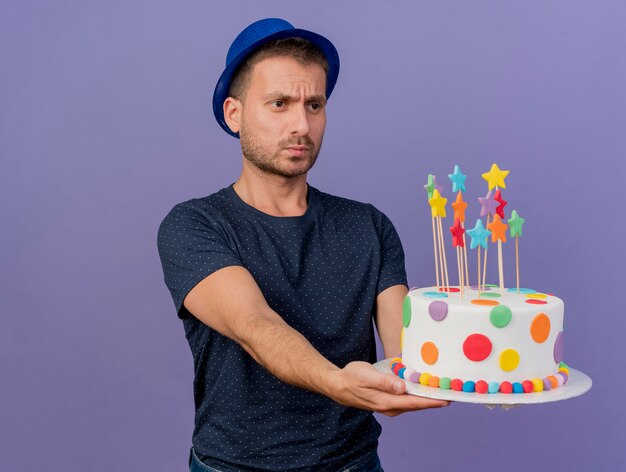 Foto gratuita l'uomo caucasico bello senza tracce che porta il cappello blu tiene e guarda la torta di compleanno isolata su fondo viola con lo spazio della copia