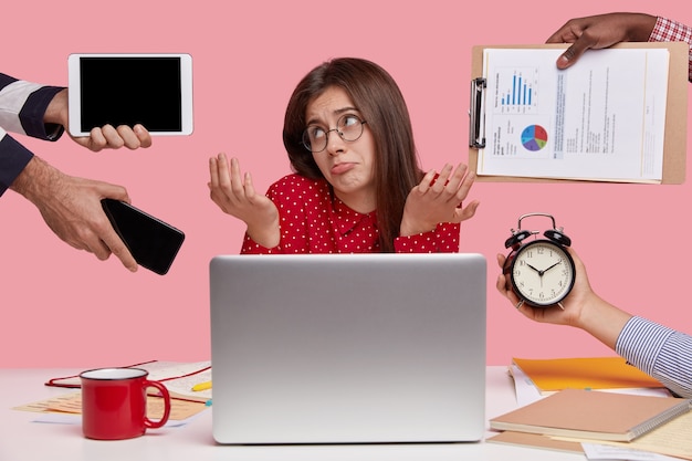 Free photo clueless indecisive female model looks in bewilderment, shruggs shoulders, surrounded with papers, modern gadgets