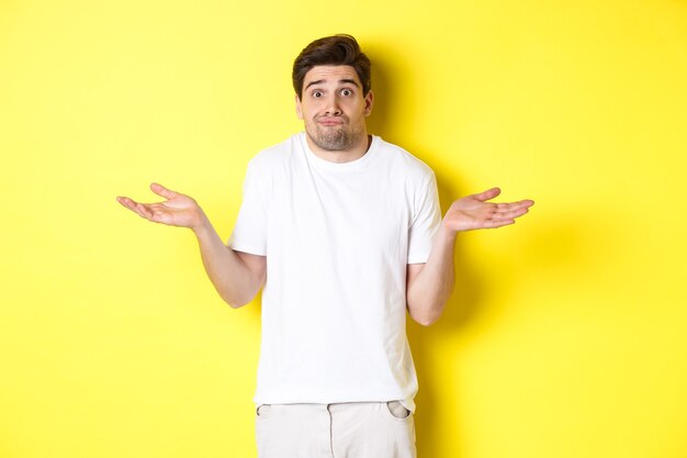 Clueless man shrugging, spread hands sideways confused, dont know anything, standing over yellow background