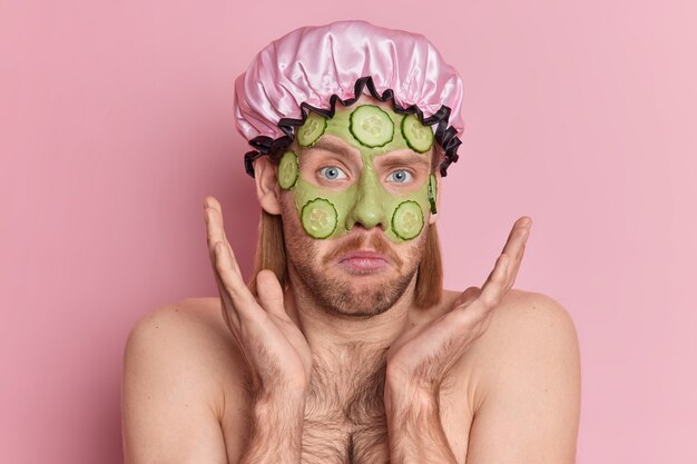 Clueless man with stubble and mustache raises palms over face feels hesitant applies green beauty mask with cucumber slices stands bare shoulders wears bath hat.