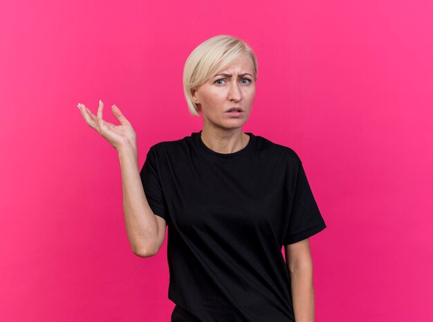 Clueless middle-aged blonde slavic woman showing empty hand looking at front isolated on pink wall with copy space