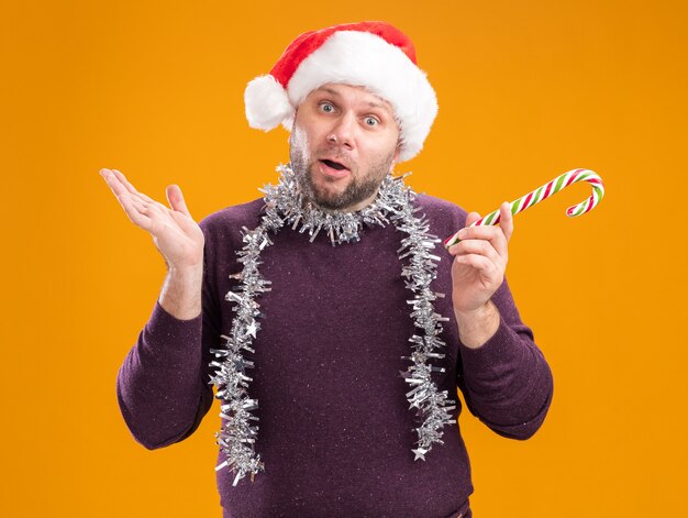 Clueless middle-aged man wearing santa hat and tinsel garland around neck holding christmas sweet cane looking at camera showing empty hand isolated on orange background