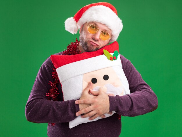 Clueless middle-aged man wearing santa hat and tinsel garland around neck with glasses holding santa claus pillow  isolated on green wall