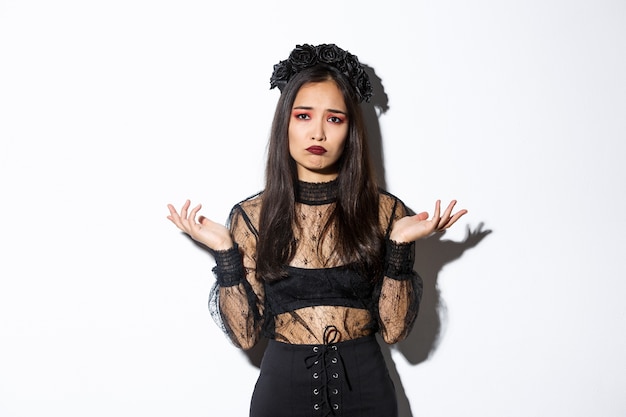 Free photo clueless pensive asian girl in witch costume looking indecisive, shrugging with hands spread sideways, looking sad over white background.