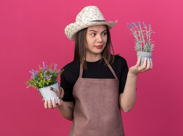 Clueless pretty caucasian female gardener wearing gardening hat holding and looking at flowerpots