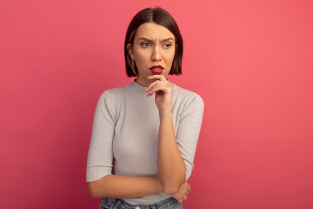 Clueless pretty caucasian woman holds chin and looks at side on pink