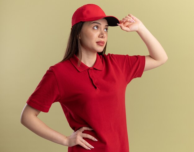 Clueless pretty delivery woman in uniform puts hand on cap and looks at side