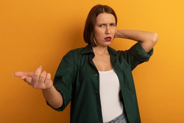 Clueless pretty woman puts hand on head behind and points at side isolated on orange wall