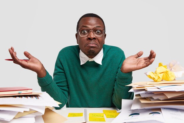 Clueless puzzled dark skinned man shruggs shoulders, feels confused as cant finish task, wears elegant green jumper, doesnt know from what to start