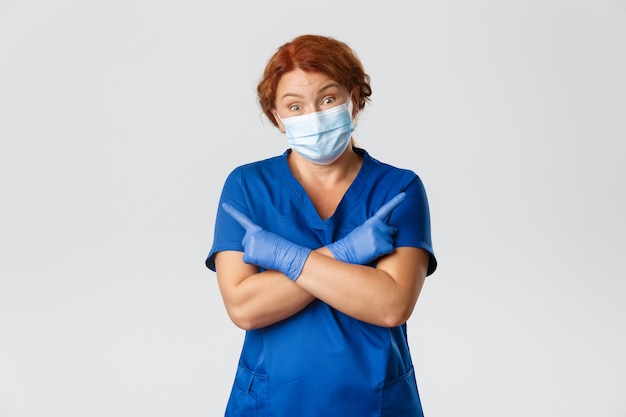 우둔한 빨간 머리 여의사, 간호사가 안면 마스크와 고무 장갑을 끼고, 옆으로 가리키며 으쓱하고 혼란스러워하고,