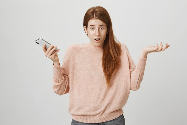 Clueless redhead woman shrugging with phone in hand