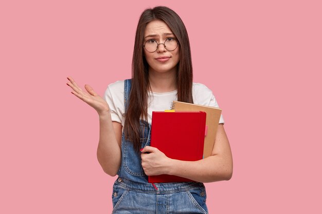 Clueless schoolgirl spreads palm, dressed in casual white t shirt and overalls, wears optical glasses, carries notepad with necessary notes