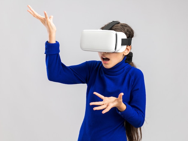 Clueless teenage girl wearing vr headset looking at side showing empty hands isolated on white wall