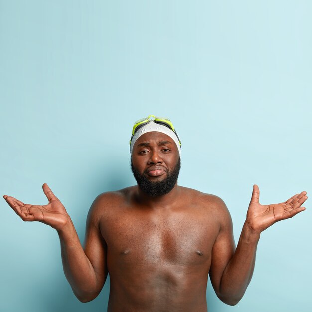 Clueless unaware swim coach spreads hands, can not decide with date of swimming competition, has training in sport centre, has dark skin, thick bristle, wears goggles on head, rubber cap, isolated