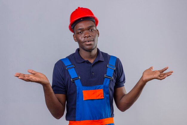 Clueless young african american builder man wearing construction uniform and safety helmet shrugging shoulders looking uncertain and confused having no answer spreading palms standing