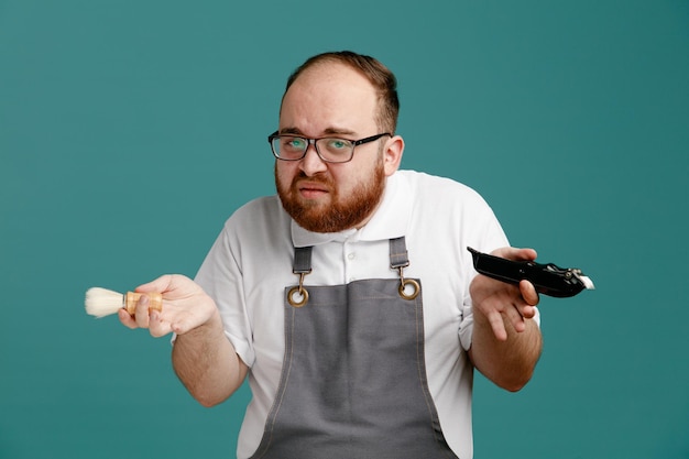 Clueless young barber wearing uniform and glasses holding shaving brush and hair trimmer looking at camera isolated on blue background