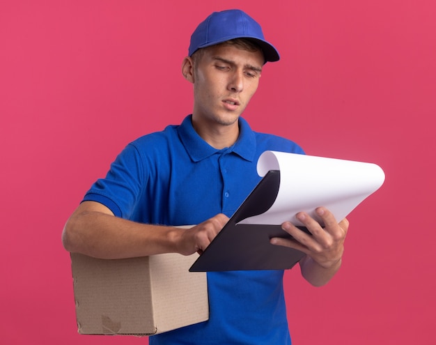 Clueless young blonde delivery boy holds cardbox and looks at clipboard