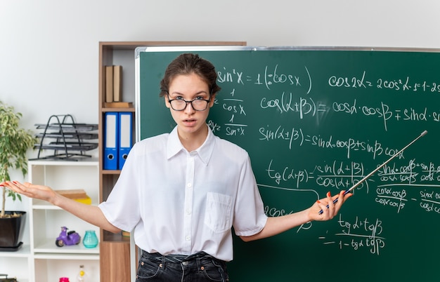 Clueless young blonde female math teacher wearing glasses standing in front of chalkboard  holding teacher pointer stick showing empty hand in classroom