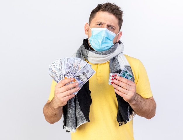 Free photo clueless young blonde ill man wearing medical mask and scarf holds money and packs of medical pills isolated on white wall