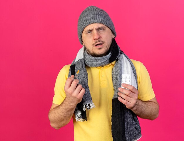 Clueless young blonde ill slavic man wearing winter hat and scarf holds syringe