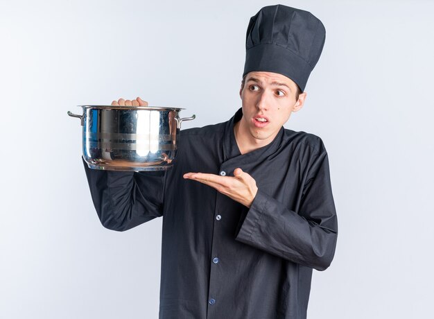 Cuoco maschio giovane biondo senza indizi in uniforme da chef e cappuccio che tiene e indica la pentola guardando il lato isolato sul muro bianco