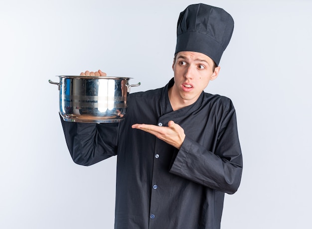 Clueless young blonde male cook in chef uniform and cap holding and pointing at pot looking at side isolated on white wall
