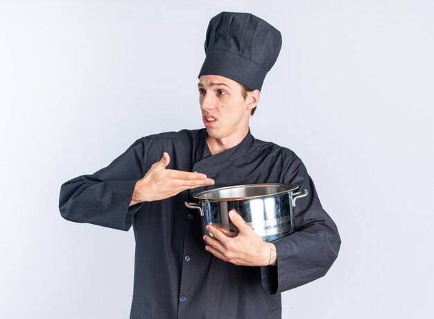 Clueless young blonde male cook in chef uniform and cap looking at side holding and pointing at pot 